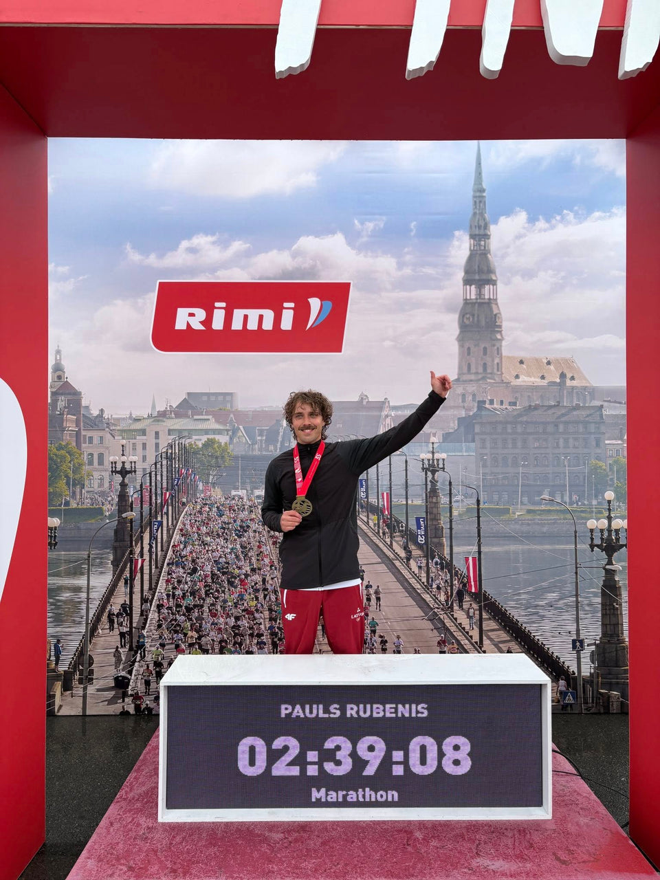 Pauls Rubenis uz Rimi Rīgas maratona goda pjedestāla ar medaļu; tablo rāda 02:39:08